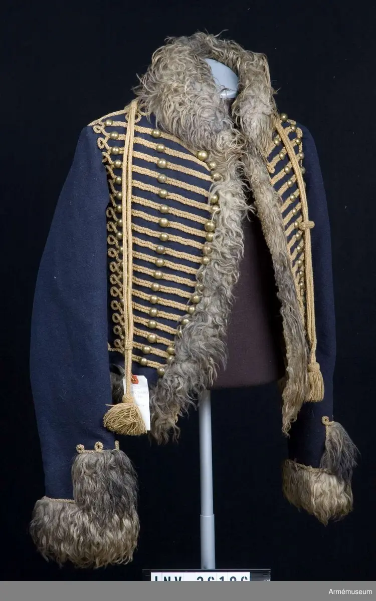 Blå päls med krage och ärmuppslag av gråbrunt fårskinn, med snören av gult ullgarn.

Pälsen är sydd av mörkblått kläde och är helfodrad med vitt, numera gulnat, korthårigt fårskinn. Ärmar, krage, framkanter och nederkant har bräm av gråbrunt fårskinn med lång lugg.
Plagget är konstruerat med fram- och sidstycken i ett samt helt bakstycke med litet skört i sidsömmarna. Ärmarna är ensömsärmar eller S-ärmar.
Tvärs över bröstet löper 18 rader snörmakerier gjorda av gul löst spunnen tvåtrådig ullsnodd. 5 knapprader, varav de två yttersta på varje sida har kullriga knappar medan knappraden i mitten som är avsedd att knäppas har klotknappar. Samtliga knappar är gjorda i en mässingslegering.
Ärmarna är kantade med päls. Pälsbrämet går upp i en "ruta" på framärmen, kantad med snodd. På baksidan av plagget nertill under ärmarna sitter två separata "rutor" av päls kantad med snodd. Sidsömmarna bak är markerade med snoddar som också pryder skörtets veck.
På baksidan av plagget nedanför kragen är det fäst ett flätat hängsle vars ändar hänger ner från varje axel och fram till plaggets främre nederkant. Varje del av hängslet är avslutat i en tofs. Vänster hängsle har ett spolformat knäppe klädd med snörmakeriarbete, medan det högra har en knut. Dessa är avsedda att hålla ihop plagget när det hänger öppet över vänster axel.
På vänster axel sitter en axelträns av dubbel snodd som sätts fast med en mässingsknapp.