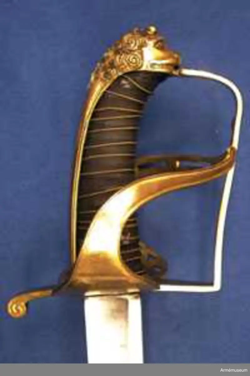 Klingans bredd upptill: 30 mm.Klingan är krökt, skålslipad och har nedtill en svart  betonad, tveegad klack. Fästet är av förgylld mässing. Kaveln är klädd med svart skinn en spiralräffla 16 varv. I bottnen på denna räffla är en av två parter hopsnodd, tunn mässingtråd anbringad. Baktill har kaveln ryggbeslag, som upptill övergår i en kappa, vilken är ciselerad i form av ett lejonhuvud och på vilken sitter en liten nitknapp. Parerstången är en rak och har fyrkantigt tvärsnitt samt av- slutas baktill av en nedböjd, på sidorna med ett spiralornament prydd knapp. Från parerstången främre ände utgår handbygeln i rät vinkel. Handbygeln är längs mitten litet försänkt och har längs var och en av framsidans kanter en ingraverad rand. Litet framför kappan genombrytes den av ett runt hål, och kring detta hål finns ciselerade ornament vilka bilda en stiliserad krans. På yttersidan finnes en uppåtböjd, delvis genombruten parerplåt, som är prydd med krönt sköld med tre kronor, vilken sköld omgives av ett par palmkvistar, allt i ciselerat arbete. Från parerplåtens spets utgår en delvis genombruten och sparsamt med ciselerade ornament prydd S-formig spång, som går till hand- bygelns övre del. På innersidan finns en liknande men mindre, ej genombruten och ej ciselerad parerplåt, från vars spets en S-formig spång går till handbygeln, vilken den träffar något längre ned än ytter- spången. Innerspången prydes längs kanterna av ingraverad rand.