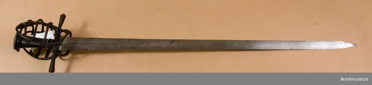 Klingan är tveeggad, har upptill på bägge sidor grund skålslipning eller bred, grund blodrand, men är i övrigt flack. Fästet är av svärtat järn och har hel gallerkorg. Knappen är låg och har upptill formen av en stympad, fyrkantig pyramid. Där- efter följer nedåt en flat 3 mm bred kant. Knappens nedre del  bildar en slags kort, bred, nedåt smalare hals med ovalt tvärsnitt. Kaveln har ovalt tvärsnitt, är vidare uppåt och är lindad med snören, som bestrukits med svart fernissa. Parerstången är rak, har kvadratiskt tvärsnitt med en kant vänd uppåt. Mot ändarna vidgas den, men ändarna har pyramidformig avslutning. Handbygelns övre ända är framåtböjd och står nära och i jämnhöjd med knappen. Framtill utgår från korset en fingerhake. På terssidan finns vid korset en stor, oval parerring. Från fingerhakens nedre ända går på terssidan en spång utåt och uppåt till parerringen, vilken den träffar strax framför dess bakre krök. Från motsvarande ställe på parerringens  övre sida går en spång något bakåt sedan utåt och uppåt, så inåt och litet nedåt till handbygelns övre del, som den träffar strax nedanför bygelns övre ända. Från den horisontala parerbygelns mitt och strax bakom dess främre krök går från vardera stället en bygel i båge uppåt till den nyss omtalade tersspången, vilken dessa byglar träffar i samma punkt ett stycke bakom dess övre ända. I övrigt utgörs skyddet på terssidan av tre horisontala spänger, som går från handbygeln till tersspången, och som tillsammans har formen av ett på bredden starkt utdraget S. På kvartsidan går från fingerhakens nedre ända en spång till parerstångens bakre arm, som den träffar strax bakom korset. Från mitten av denna spång går en annan längre spång något bakåt, sedan utåt och uppåt, så framåt och slutligen nedåt till handbygeln, som den träffar strax nedanför bygelns övre ända. Strax bakom den främre kröken på den förstnämnda kvartspången går en bygel i båge uppåt till den sistnämnda spången, som den träffar en bit bakom dess övre ända. I övrigt utgörs skyddet på kvartsidan av tre horisontala spänger, som går från handbygeln till den längre kvartspången. Dessa tre sistnämnda kvartspänger har tillsammans formen av ett på bredden starkt utdraget S. Alla byglar och spänger har cirkelrunt tvärsnitt.