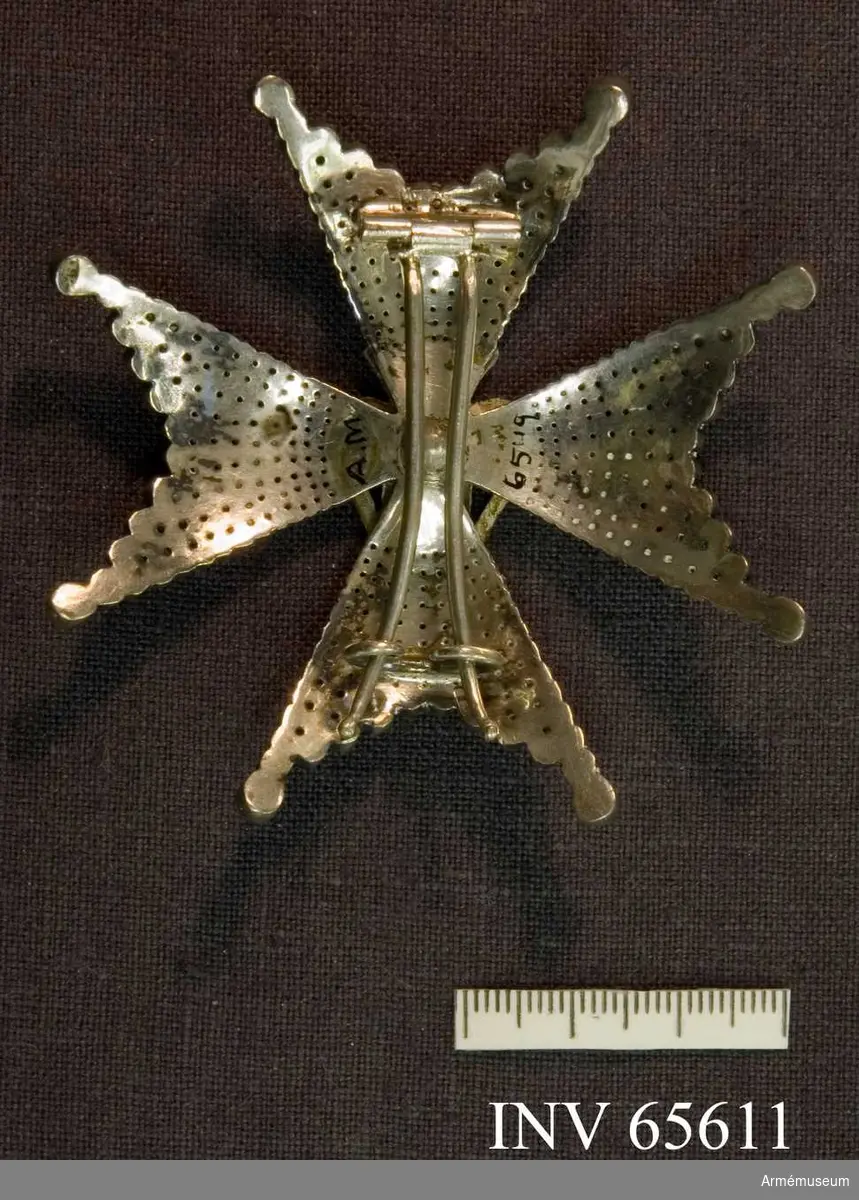 Grupp M II.   

Ett johannitkors av fasetterade silverpärlor med kluvna armar och uddarna slutande i en fasetterade kulor. 
Ovanpå korset i upphöjt arbete en stiliserad sädeskärve (vase) samt en kunglig krona, allt i silver.   