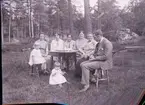 En familj sitter vid ett bord i en trädgård. 
Fyra kvinnor i olika åldrar, en man och två små barn. 

Uppgifter av HBF: 