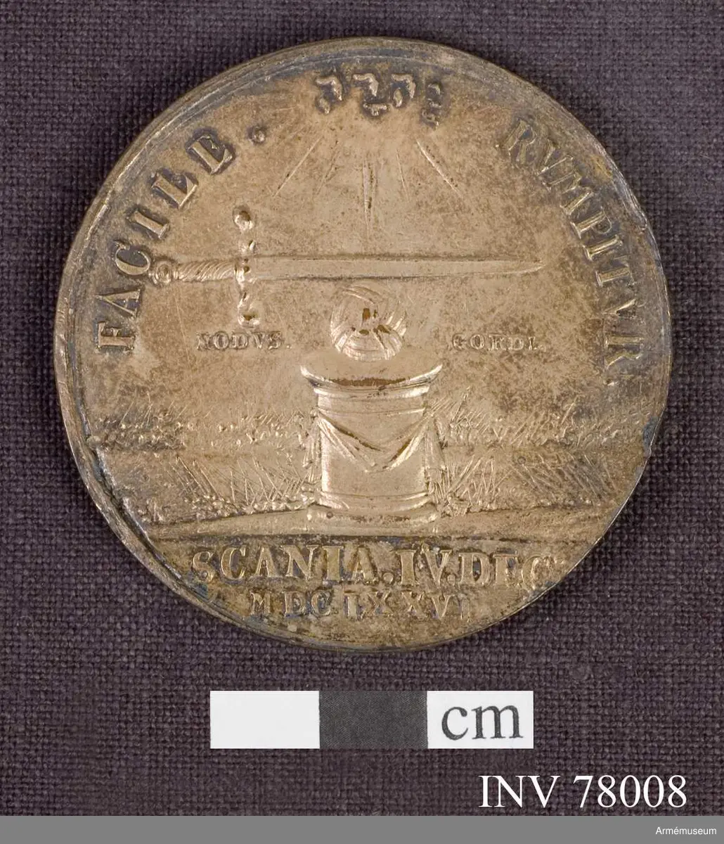 Grupp: M. 
Åtsidan: CAROL. XI. D G. REX. SVEC: Bröstbild åt höger, i harnesk med ett uppvänt ansikte på övra delen, samt harnesk med ett högt Medusahuvud på axeln och mantelveck - Under armen B.

Frånsidan: FACILE RVMPITVR. Ett blottat svärd svävar horisontelt över den sk gordiska knuten, ett av tåg konstigt hopknutet klot, som ligger på ett runt altare. Vid sidorna av knuten: NODVS. GORDI. I bakgrunden ses de stridande härarna, den danska flyende. Överst ett strålande JHVH (med hebreiska tecken). I avskärningen: SCANIA. IV. DEC. M.DC. LXXVI. på två rader.