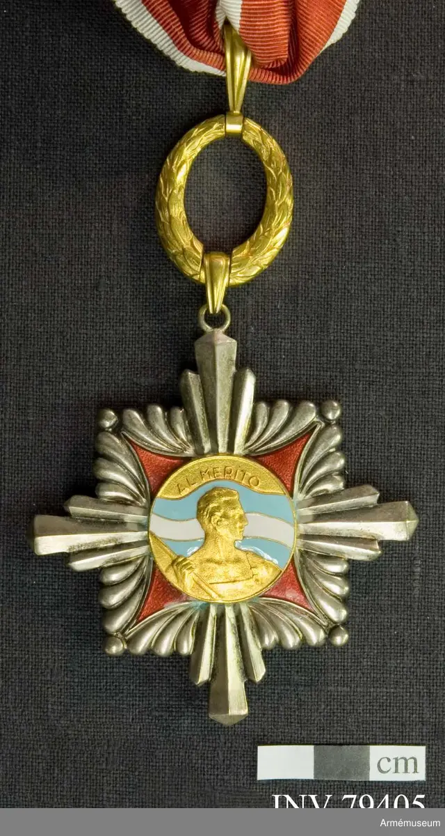 Kommendörstecken av argentinsk förtjänsteorden. På framsidan en man hållande argentinas fana i blått och vitt med texten "AL MERITO". På baksidan vapenskölden för Argentinas president.

Med band i rött med vita kanter.