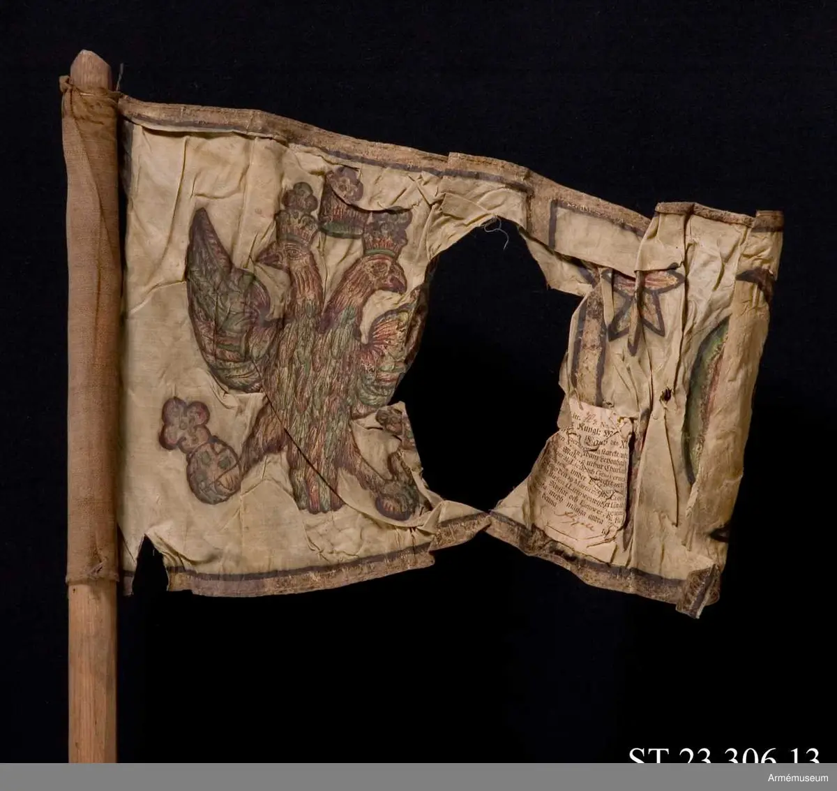 Duk av vit sidentaft och strumpa av rött linne. Motivet målat i guld- och silver samt rött och grönt bestående av en krönt örn, måne, sol och stjärnor, På duken en pappersetikett som anger att den tagits i slaget vid Saladen 1703.