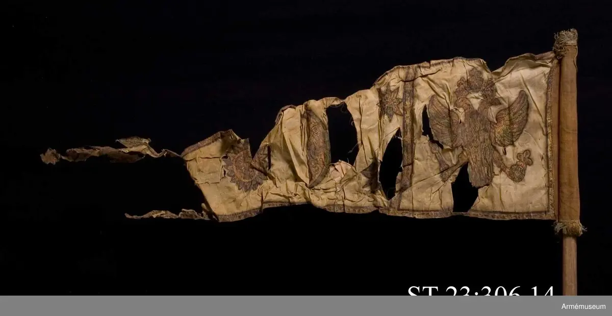 Duk av vit sidentaft och strumpa av rött linne försedd med frans vid öppningarna. Motivet målat i guld- och silver samt rött och grönt bestående av en krönt örn, måne, sol och stjärnor, På duken en pappersetikett som anger att den tagits i slaget vid Saladen 1703.