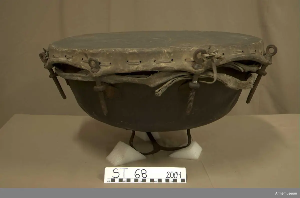 Kittel av koppar och skinn av kalv med fast spännring. Tredelad fot av järn. 7 skruvar och skruvfästen av järn. Bärrem av kalvskinn i två lager med spänne av järn.