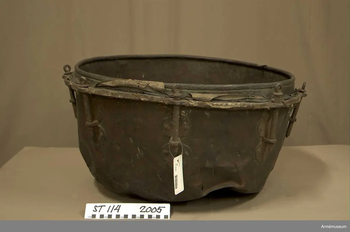 Tillstånd 2005:
Kittel: kraftigt bucklig och sprucken på flera ställen. En mängd stickmärken i storlek 1 cm. - 4 cm. Fot: saknas. Spännskinn: saknas, förutom en liten flik - hängandes över kanten in i pukan - likaså är skinnet lämnat längsmed kittelkanten fastsytt genom ett smalt läderband. Spännring: rostig. Spännskruv: 8 st. Skruvfästen: 8 st. Bärrem: saknas.