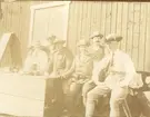 Män i Landstormsuniform sitter på en bänk framför en träbyggnad i Löttinge, Täby, 1914. På bordet framför står flaskor och koppar.