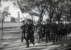 Fanvakt paraderar vid uppvisning på Arméns underofficersskola (Försvarets läroverk) i Uppsala 1949. I bakgrunden syns järnvägen och bostadshus.