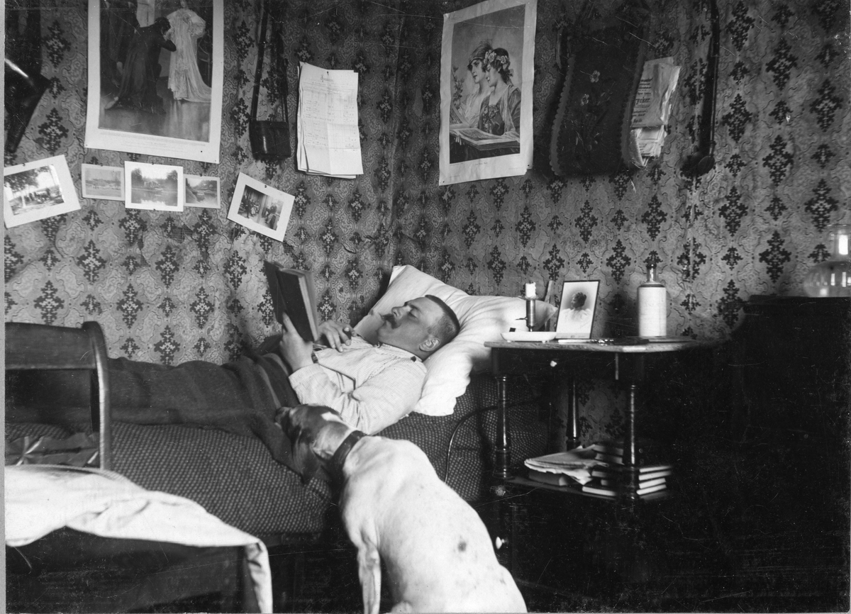 Officer vid Älvsborgs regemente ligger i sängen och läser en bok och röker en cigarr. På golvet nedanför sitter en hund.