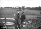 En man och en kvinna vid en gärdesgård. En gård med omgivande fält syns i bakgrunden.