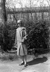 En kvinna klädd i kappa och hatt står på en väg framför en häck. I bakgrunden skymtar ett stort hus.