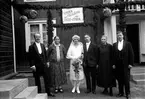 Ett brudpar med föräldrarna uppställda för fotografering av Gustav Andersson, Jönköping, framför en granrisklädd veranda. Där hänger också papperslyktor och en skylt med texten: Lycka och välgång för Olle + Ebba.