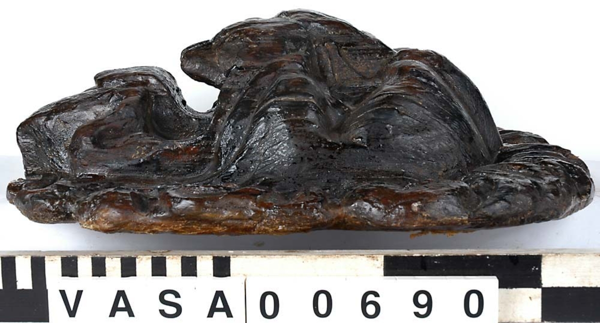 Skulpturdel, utgör större delen av en lejonmaskaron. I sin helhet är lejonmaskaronen rund, eller obetydligt rektangulär till formen, och utförd i två sektioner. Ansiktet har stora, runda ögon med kraftiga ögonbrynsbågar. Nosen är platt och bred och klart avgränsad från den rundade överkäken. Det vidöppna gapet har breda, något inåtsvängda tänder och en bred tunga. Underkäken är försedd med två strutformade hårtofsar. Lejonets man är i det närmaste jämnbred och kraftig.

Skulpturen är välbevarad, dock något ytsliten.



Text in English: Part of a sculpture, the larger part of a lion mask. The lion mask is in its entirety round or slightly rectangular in form, and made in two sections. The lion's face has large round eyes and powerful eyebrows. The flat broad nose is clearly separated from the rounded upper jaw. The jaws are wide open with broad and somewhat concave teeth and a broad tongue. The lower jaw has two cone-shaped tufts of hair. The lion''s mane is powerful and almost of even width.

The sculpture is well preserved but worn on the surface.