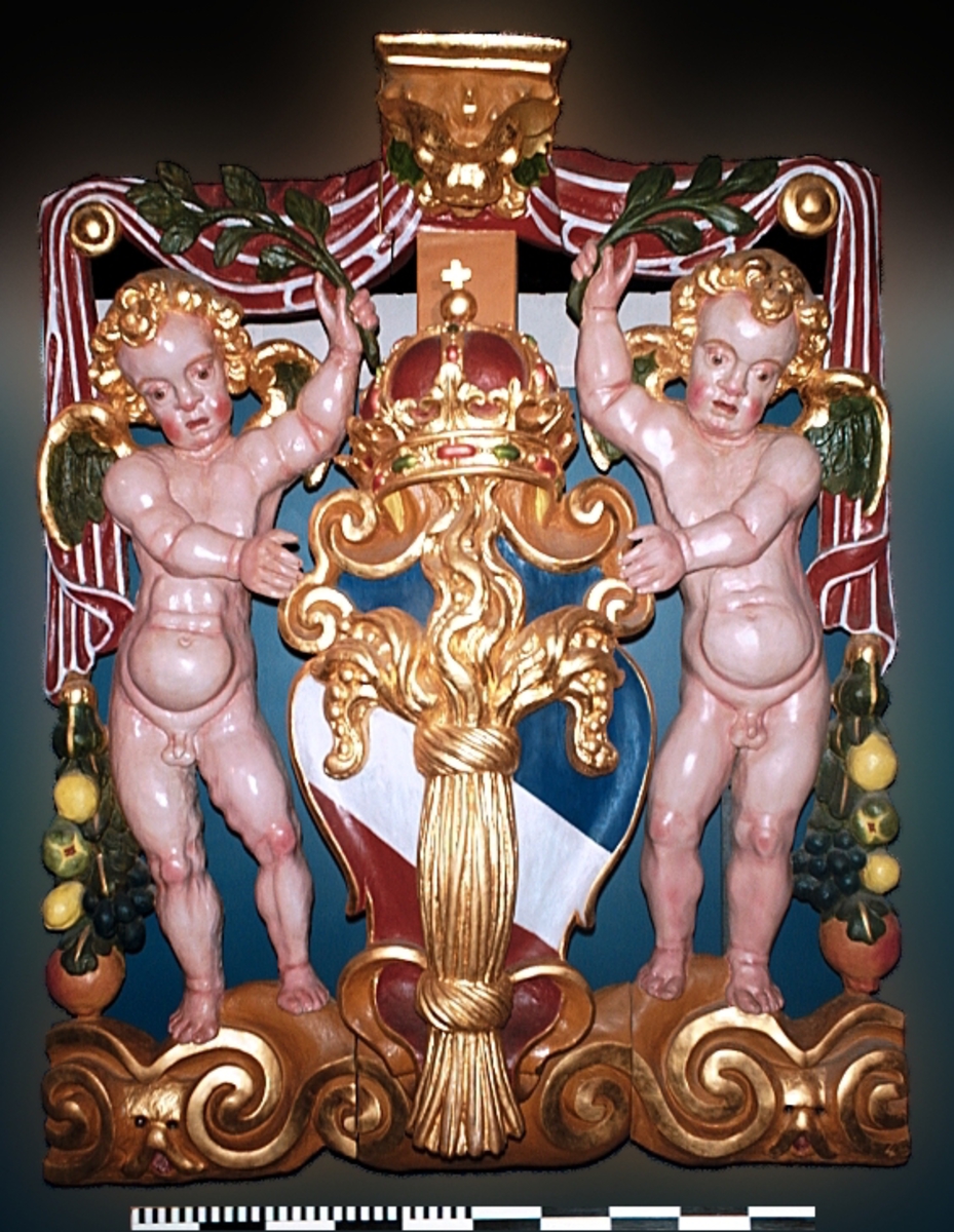 Skulptur av en putto stående på postament med omgivande ornamentik.
Putton har ett frodigt och undersätsigt utseende, med avrundade former. Puttons vänstra hand vilar på en sköld, medan höger hand är uppåtsträckt, hållande en olivkvist.
Ovanför putton sitter ett veckrikt draperi, "upphängt" på en stång med profilerad ände. Där det nedhängande draperiet slutar vidtar tunga fruktfestoner som når ned till postamentet, vilket är dekorerat med symmetriskt ställda rullverksspiraler. Rullverksspiralerna är utformade så att de bildar krumma vädurs- eller gumsehorn till ett i postamentet utsnidat djurhuvud.
Puttons ansikte, vänster arm samt mage är snidade separat. Skulpturens baksida är slät.

Text in English: Sculpture of a putto, standing on a base surrounded by ornamentation.
The exuberant plump cherub has a well-rounded body. Right hand rests on a shield. The left hand holds an olive branch aloft.

Above the putto''s head hangs a heavily-folded drapery. The drapery forms a swag as it hangs from a curtain rod. The prominent ends of the curtain rod are carved in relief. Heavy festoons of fruit flow from the drapery ends to the feet of the putto. The symmetrical scrollwork on the base takes on the appearance of ram''s horns when combined with the imprecise representations of animal heads.
Face, left arm and belly are carved separately. Reverse side is smooth.