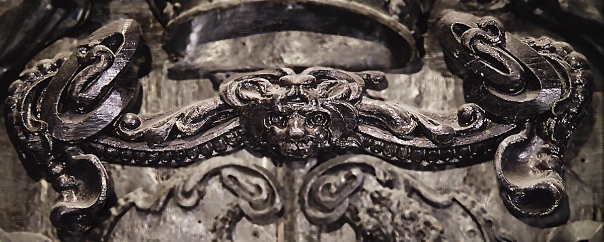 Broskornament. Ornamentet är utformat som ett kattdjursliknande huvud från vilket utgår "armar", dekorerade med blad och broskvoluter. Ändpartierna är förtjockade med elipsformade broskvoluter, blad och nedhängande flikar. Baksidan är slät.
Ornamentet är brutet på baksidans mittparti.

Text in English: Earshell ornament consisting of a cat-like head, from which extend "arms" decorated with ellipse-shaped pearlwork scroll leaves and downward-hanging leaf-blades with thickened ends. The reverse side is smooth.
The ornament is broken in the middle on the reverse side.