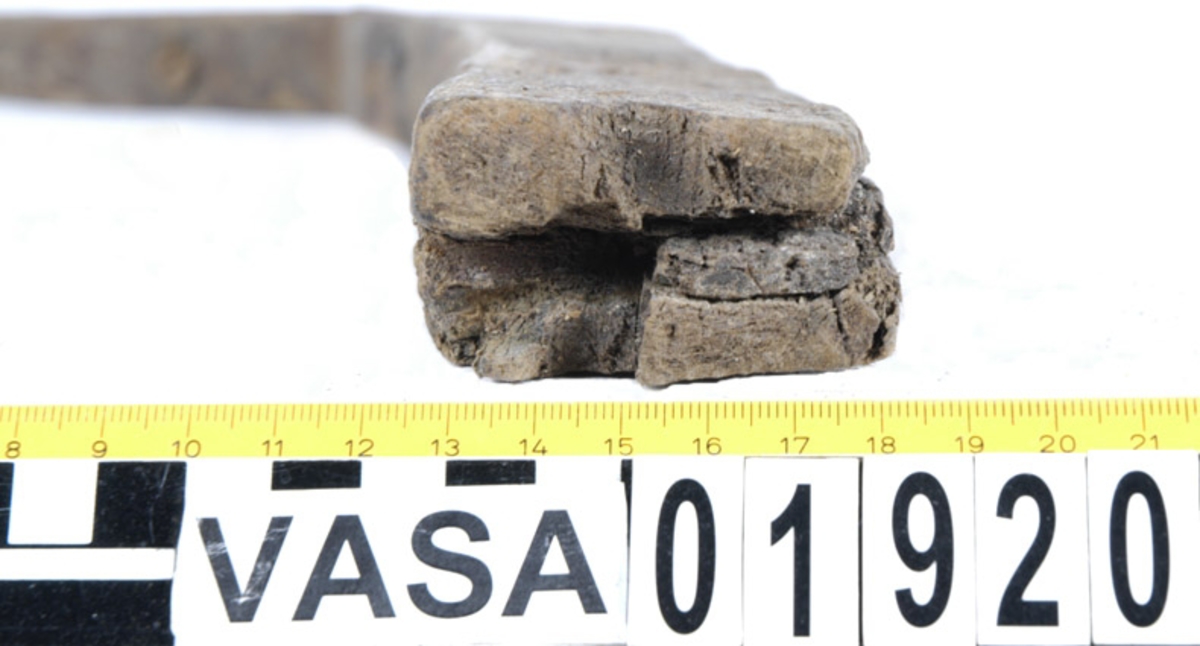 Stäv. Stäv till mindre båt. Stävtoppen verkar sluta i en intappad stumlask. Flera spikhål i det svagt konkava område som har utgjort anläggningsyta för bordgångarna. Ingen spunning. Stävdelen har flera grova, i längsled, genomgående spikhål (6 mm i fyrkant) som uppvisar avtryck efter skalle eller bricka på insidan. Vid krit där stävdelen övergår i köldel finns en klack som troligen har berett plats för en järnskoning av stäven. Inga rostspår av denna finns dock. Det som utgör köldelens underkant bär spår av flera, ej genomgående, spikar som sannolikt också har varit infästning för en slags skoning. Köldelen har varit infäst i en överliggande kölplanka och utgjort den främsta (eller akterligaste) vertikala delen i en T-köls konstruktion. Två 18 mm dymlingar har fäst den till kölplankan. Den ena är bevarad men avbruten, den andra indikeras av ett halvt dymlinghål i den avbrutna änden av köldelen.