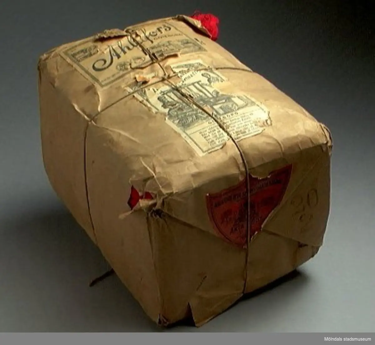 En förpackning med rött bomullsgarn (äkta rödfärg nr 39). Garnmängden (2 kg?) paketerat i brunt omslagspapper, med snöre runt om. Tre olika etiketter från Ahlafors är påklistrade. Pappret är trasigt.Tidigare sakord: garn i förpackning.