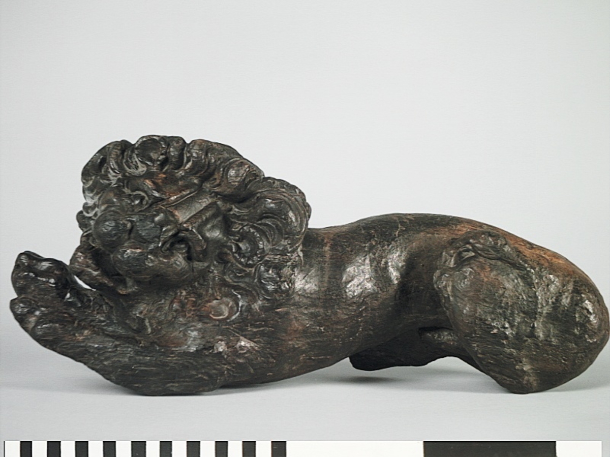 Skulptur av ett litet, liggande lejon med utsträckta framben och hopdragna bakben.
Lejonet är riktat mot vänster, sett från betraktaren. Det snett hållna huvudet återges i det närmaste i en face. Större delen av svansen saknas. Den befintliga delen av svansen avtecknar sig mot lejonets vänstra lår.

Lejonet är endast delvist rundskulpterat. På baksidan, i höjd med huvudet och höger framben, återfinns ett stort, snett urtag som bildar en slät anliggningsyta. Skulpturens framparti är således endast till hälften så tjockt som dess bakparti.
Bakkroppen är kraftigt sliten och skadad. Vänster bakben saknas, liksom större delen av svansen.

Text in English: Sculpture of a small reclining lion with its forelegs extended and its  hind legs tucked beneath its body.
Facing to the observer''s left, the head, held crookedly, is nearly en face. A large part of the tail is missing, and the remainder curves towards the left hind leg.

The lion is only partly three-dimensional. On the rear at the head height and at the right foreleg a large oblique hole forms a flat area for attachement, thus the forepart of the sculpture is thus only half as thick as its rear.
The rear of the body is badly worn and damaged. The left hind leg has been broken off.