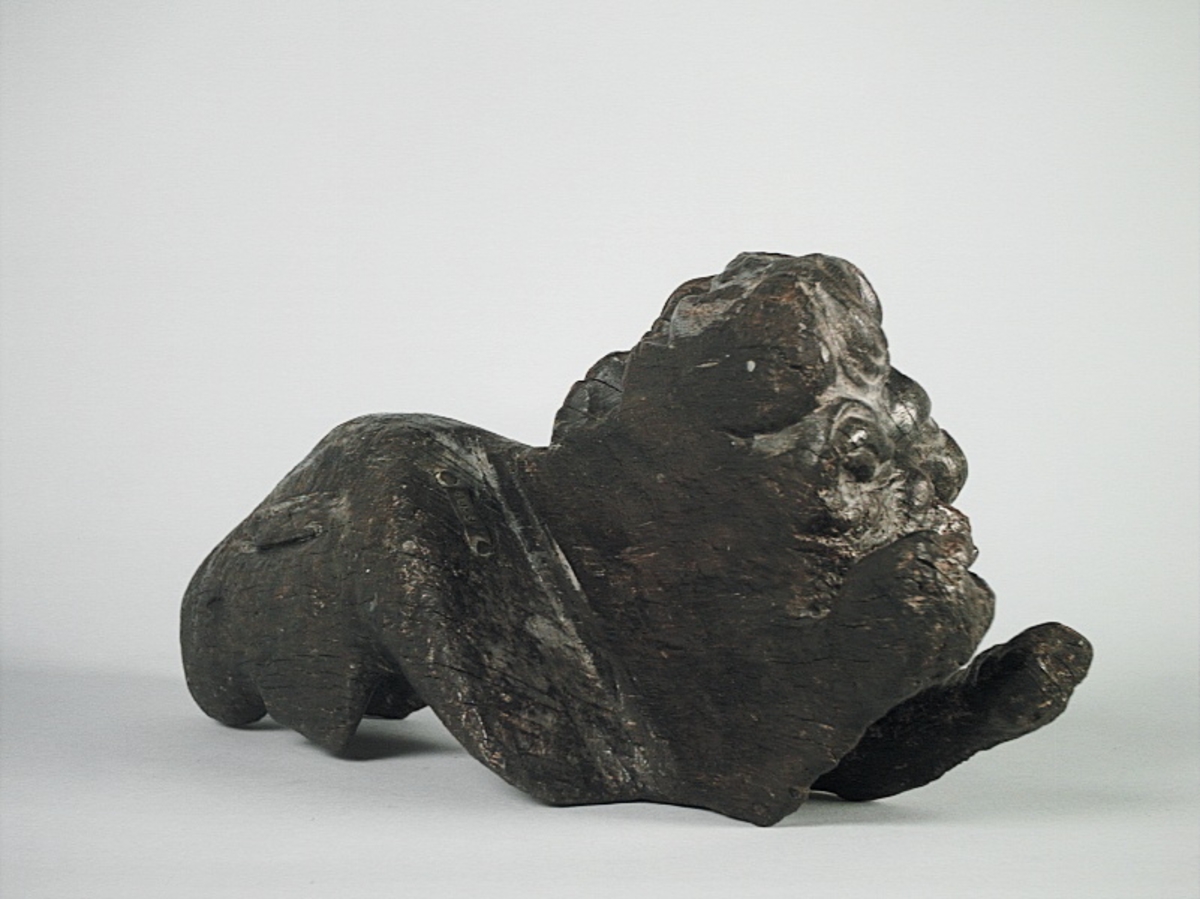 Skulptur av ett litet, liggande lejon med utsträckta framben och hopdragna bakben.
Lejonet är riktat mot vänster, sett från betraktaren. Det snett hållna huvudet återges i det närmaste i en face. Större delen av svansen saknas. Den befintliga delen av svansen avtecknar sig mot lejonets vänstra lår.

Lejonet är endast delvist rundskulpterat. På baksidan, i höjd med huvudet och höger framben, återfinns ett stort, snett urtag som bildar en slät anliggningsyta. Skulpturens framparti är således endast till hälften så tjockt som dess bakparti.
Bakkroppen är kraftigt sliten och skadad. Vänster bakben saknas, liksom större delen av svansen.

Text in English: Sculpture of a small reclining lion with its forelegs extended and its  hind legs tucked beneath its body.
Facing to the observer''s left, the head, held crookedly, is nearly en face. A large part of the tail is missing, and the remainder curves towards the left hind leg.

The lion is only partly three-dimensional. On the rear at the head height and at the right foreleg a large oblique hole forms a flat area for attachement, thus the forepart of the sculpture is thus only half as thick as its rear.
The rear of the body is badly worn and damaged. The left hind leg has been broken off.