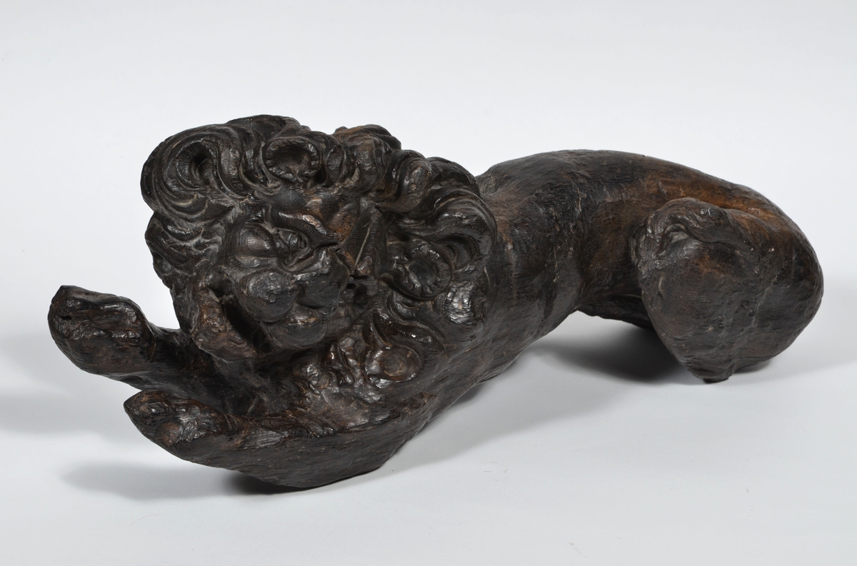 Skulptur av ett litet, liggande lejon med utsträckta framben och hopdragna bakben.
Lejonet är riktat mot vänster, sett från betraktaren. Det snett hållna huvudet återges i det närmaste i en face. Större delen av svansen saknas. Den befintliga delen av svansen avtecknar sig mot lejonets vänstra lår.

Lejonet är endast delvist rundskulpterat. På baksidan, i höjd med huvudet och höger framben, återfinns ett stort, snett urtag som bildar en slät anliggningsyta. Skulpturens framparti är således endast till hälften så tjockt som dess bakparti.
Bakkroppen är kraftigt sliten och skadad. Vänster bakben saknas, liksom större delen av svansen.

Text in English: Sculpture of a small reclining lion with its forelegs extended and its  hind legs tucked beneath its body.
Facing to the observer''s left, the head, held crookedly, is nearly en face. A large part of the tail is missing, and the remainder curves towards the left hind leg.

The lion is only partly three-dimensional. On the rear at the head height and at the right foreleg a large oblique hole forms a flat area for attachement, thus the forepart of the sculpture is thus only half as thick as its rear.
The rear of the body is badly worn and damaged. The left hind leg has been broken off.
