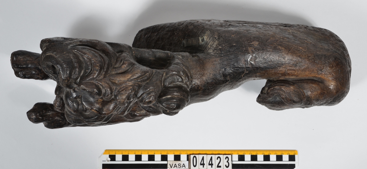 Skulptur av ett litet, liggande lejon med utsträckta framben och hopdragna bakben.
Lejonet är riktat mot vänster, sett från betraktaren. Det snett hållna huvudet återges i det närmaste i en face. Större delen av svansen saknas. Den befintliga delen av svansen avtecknar sig mot lejonets vänstra lår.

Lejonet är endast delvist rundskulpterat. På baksidan, i höjd med huvudet och höger framben, återfinns ett stort, snett urtag som bildar en slät anliggningsyta. Skulpturens framparti är således endast till hälften så tjockt som dess bakparti.
Bakkroppen är kraftigt sliten och skadad. Vänster bakben saknas, liksom större delen av svansen.

Text in English: Sculpture of a small reclining lion with its forelegs extended and its  hind legs tucked beneath its body.
Facing to the observer''s left, the head, held crookedly, is nearly en face. A large part of the tail is missing, and the remainder curves towards the left hind leg.

The lion is only partly three-dimensional. On the rear at the head height and at the right foreleg a large oblique hole forms a flat area for attachement, thus the forepart of the sculpture is thus only half as thick as its rear.
The rear of the body is badly worn and damaged. The left hind leg has been broken off.