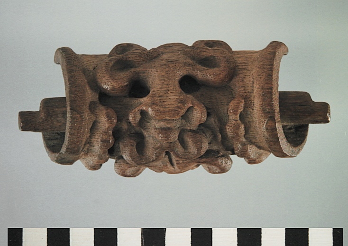 Rullverksornament.
Skulpterat litet, spänneliknande ornament, dekorerat i broskverksstil.
Det långsmala ornamentet är format som ett stycke kraftigt upprullad bit näver eller läder, med konkav bak- eller undersida. På baksidan är ornamentet profilerat för en list. Broskdekoren på ovansidan är formad som en maskaron.
Ornamentet är mycket välbevarat.

Text in English: A small, buckle-like ornament, decorated in a style similar to earshell work.
The long narrow ornament is formed as a piece of tightly rolled bark or leather, with a concave back or underside so as to match a batten or the like. The reverse side is profiled to fit a moulding. The earshell work on the upper side is shaped as a mask.
The ornament is well preserved.