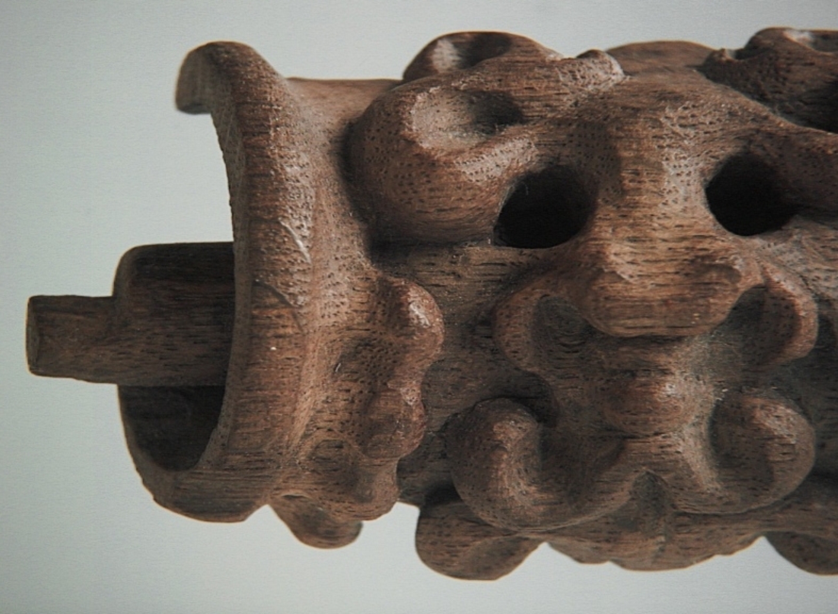 Rullverksornament.
Skulpterat litet, spänneliknande ornament, dekorerat i broskverksstil.
Det långsmala ornamentet är format som ett stycke kraftigt upprullad bit näver eller läder, med konkav bak- eller undersida. På baksidan är ornamentet profilerat för en list. Broskdekoren på ovansidan är formad som en maskaron.
Ornamentet är mycket välbevarat.

Text in English: A small, buckle-like ornament, decorated in a style similar to earshell work.
The long narrow ornament is formed as a piece of tightly rolled bark or leather, with a concave back or underside so as to match a batten or the like. The reverse side is profiled to fit a moulding. The earshell work on the upper side is shaped as a mask.
The ornament is well preserved.