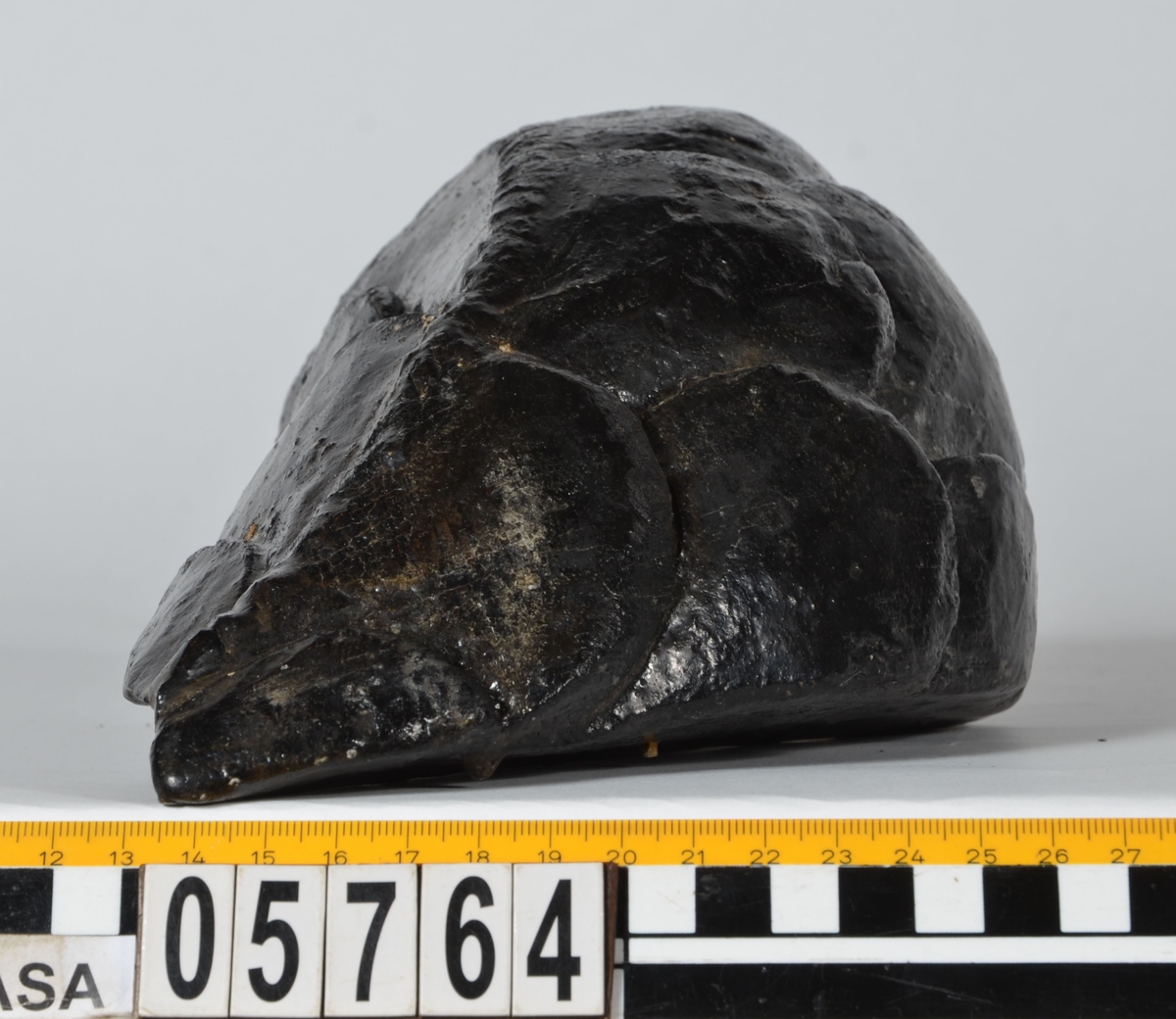 Del av skulptur, nedre del till fiskstjärt. Avsmalnande i ena änden. Rundad framsida med utsnidade fiskfjäll. Baksidan är bruten.