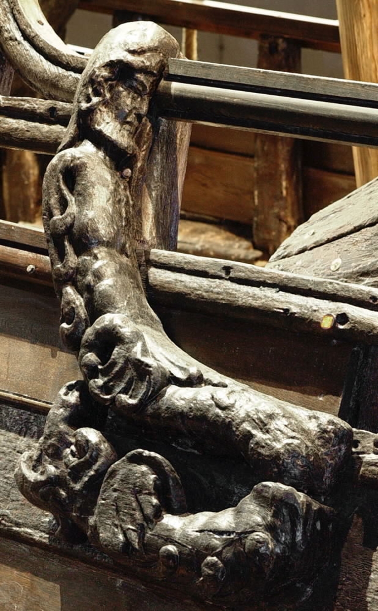 Skulptur föreställande en triton, svängd mot vänster och framställd i höger profil.
Tritonen har skäggigt ansikte, stor näsa samt en bladsmyckad hjälm på huvudet. Armarna är ersatta av långa, flikiga bladslingor. Bålens övergång i den fjällförsedda fiskstjärten markeras av stora bladbildningar. Figuren avtecknar sig i hög relief mot det släta bakstycket. Nedtill ses en fiskliknande stjärt med drakfena, vilken är fortsättningen på en under liggande draken, se fyndnummer 00628. På baksidan finns urtag för relingslisten.
Skulpturen är välbevarad.

Text in English: A sculpture of a Triton curved to the left, in right profile.
Bearded face with a long nose. Leaf-decorated helmet. The arms are replaced by long, lobed leaf formations. The transition between the male torso and the scaled fish-tail is decorated with large leaves. The figure is seen in high relief against a plain back board. Below the figure can be seen a section of another fish-like tail but actually a dragon''s fin, belonging to the dragon situated underneath, see No. 00628. There are notches on the back of the sculpture that fit into the railing.
The sculpture is well preserved.
