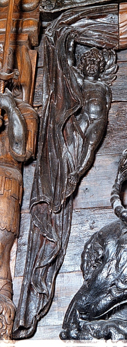 Putto samt större delen av ett draperi. Draperiet är kilformat med rak ytterkontur, och hålls undan av den svävande putton. Skulpturens baksida är slät.

Text in English: Putto and the larger part of a drapery. The drapery is wedge-shaped with straight outer contour, and is held back by the hovering putto. The reverse side is smooth.