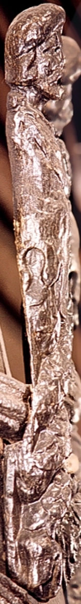 Skulpterad Triton med tjock lagerkrans på huvudet.
Figuren har stora yviga mustascher och långt, lockigt hår. Snett över bröstet är en klädnad draperad. Klädnaden är hopfäst på vänster axel.

Övergången till de snodda fiskstjärtarna döljs av ett stort, flikigt blad vars mittflik är förtjockad och upprullad. Skulpturens fotända är tapp- eller kilformad och tillplattad från sidorna. Tritonens huvud är framställt som rundskulptur. Från axlarna och nedåt är baksidan slät och plan. I höjd med övergången till stjärtpartiet finns ett kraftigt urtag. Nedtill bryter baksidans fotända av inåt.

Text in English: Sculpted Triton with thick laurel wreath on its head.
The figure has a large bushy moustache and long curly hair. A garment is draped across the chest and fastened on the left shoulder.

The transition between the upper body and fish tails is concealed by a large leaf, the middle segment of which is thickened and rolled up. The foot end of the sculpture has a flat end, seen from the side, and is wedge-shaped. The Triton''s head is carved as a three-dimensional sculpture. The rear side is smooth. There is a deep groove on the rear side at the transistion point between the male torso and the fish tails.