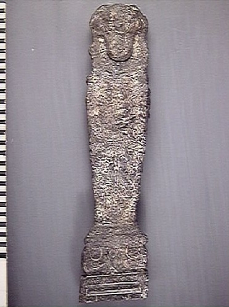 Liten skulpterad pilaster, utformad som en karyatid eller möjligen en putto. Figuren har långt hår och ett knubbigt ansikte, omgivet av en bred krage. Pilasterskaftet är dekorerat med akantusblad. Nedtill avslutas pilastern av en rikt profilerad och dekorerad sockel. Skulpturens baksida är slät.
Skulpturen är mycket sliten.

Text in English: A small carved pilaster, with a caryatid or probably a putto. The figure has a chubby face, long hair and a broad collar. The lower part of the pilaster is decorated with acanthus leaves. The figure stands on a richly profiled and decorated socle. The back of the sculpture is smooth.
The sculpture is very worn.