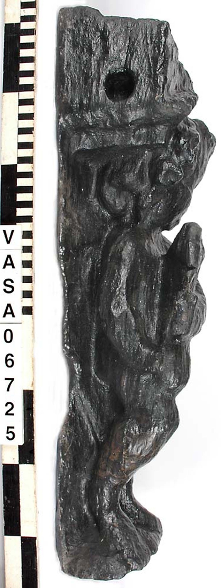 Skulptur föreställande en barnfigur spelande på en säckpipa.
Huvudet är vridet åt vänster och något nerböjt. Säckpipan hålls med bägge händerna och stöder dessutom på höger ben som är böjt. Kroppstyngden vilar på vänster ben.
Ovanför hjässan ses en snedställd äggstavslist. Det triangelformade partiet ovanför listen har utgjort anliggningsyta. Figuren står på ett minimalt underlag som troligen avser att föreställa ett stycke naturlig mark. Skulpturen har en slät baksida.
Skulpturen är kraftigt sliten och ytnött.

Text in English: This is a sculpture of a child playing a set of bagpipes.
The bagpipes are held in both hands and are further supported on the right leg, which is bent. The left leg is straight and takes the weight of the body. The head is turned slightly to the right and a trifle bent. The figure is partly draped in a cloak which conceals his right side.
Above the head the backboard comes forward at right angles. The front of the board is wide and is decorated with an egg-moulding design  which is placed diagonally across it, with the left-hand side lower than the right hand side when viewed from the front.. The figure stands upon a small flat plinth. The back of the sculpture is smooth.
The sculpture is badly worn.