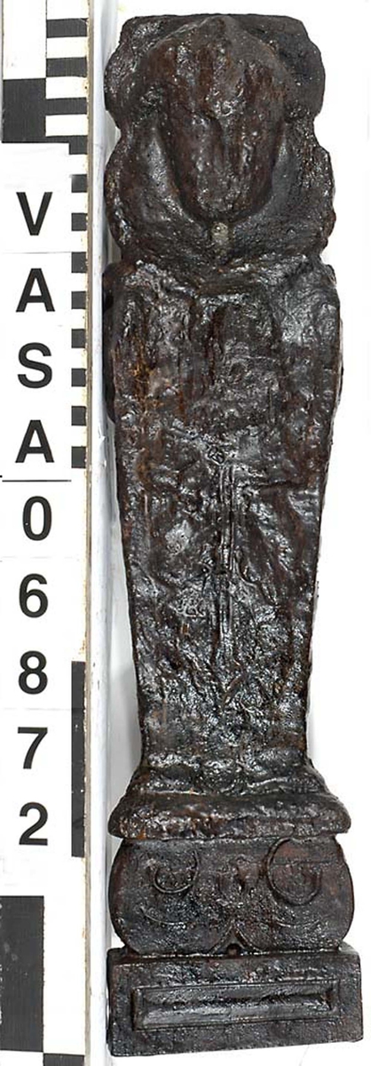 Liten skulpterad pilaster, troligtvis utformad som en karyatid eller möjligen en putto. Figuren har långt hår och ett knubbigt ansikte, omgivet av en bred krage. Det nedåt avsmalnande pilasterskaftet är dekorerat med akantusblad. Nedtill avslutas pilastern av en rikt profilerad och dekorerad sockel. Skulpturens baksida är slät.

Skulpturen är sliten, pilasterns nedre del är dock mindre sliten än den övre.



Text in English: A small carved pilaster, with a caryatid or probably a putto. The figure has a chubby face, long hair and a broad collar. The lower part of the pilaster is decorated with acanthus leaves. The caryatid stands on a richly profiled and decorated socle. The back of the sculpture is smooth.

The sculpture is worn, the lower end less so than the upper.