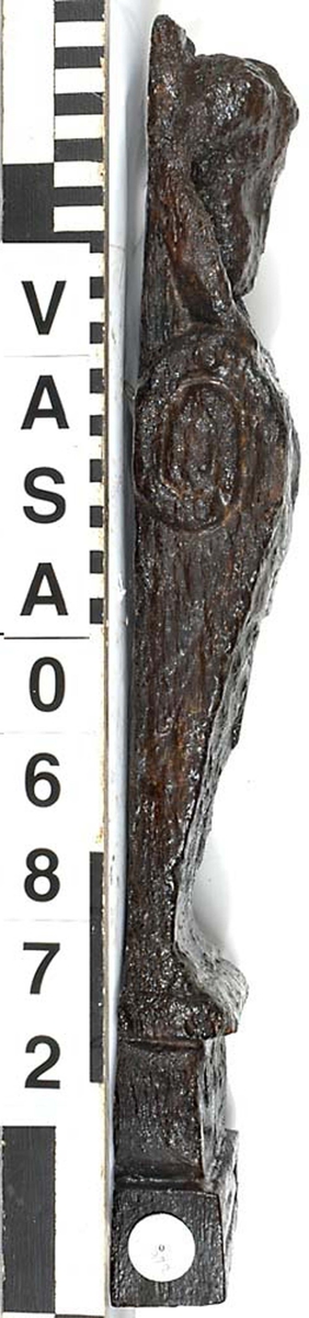 Liten skulpterad pilaster, troligtvis utformad som en karyatid eller möjligen en putto. Figuren har långt hår och ett knubbigt ansikte, omgivet av en bred krage. Det nedåt avsmalnande pilasterskaftet är dekorerat med akantusblad. Nedtill avslutas pilastern av en rikt profilerad och dekorerad sockel. Skulpturens baksida är slät.

Skulpturen är sliten, pilasterns nedre del är dock mindre sliten än den övre.



Text in English: A small carved pilaster, with a caryatid or probably a putto. The figure has a chubby face, long hair and a broad collar. The lower part of the pilaster is decorated with acanthus leaves. The caryatid stands on a richly profiled and decorated socle. The back of the sculpture is smooth.

The sculpture is worn, the lower end less so than the upper.
