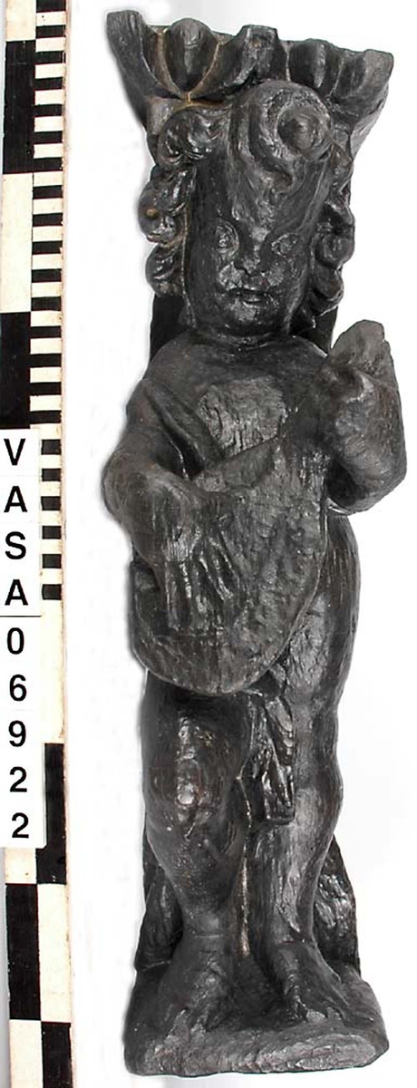 Skulptur föreställande en barnfigur med en cister (ett stränginstrument som tillhör familjen lutor).

Instrumentet hålls med bägge händer framför kroppen. Huvudet något vridet åt vänster. Kroppstyngden vilar på vänster ben medan höger ben är böjt. Utmed höger sida skymtar en mantel. Figuren står på ett minimalt underlag som troligen avser att föreställa ett stycke naturlig mark. Skulpturens baksida är slät.

Skulpturen är välbevarad.



Text in English: This is a sculpture of a child playing a cister.

It holds the instrument in both hands in front of its body. The head is turned slightly to the left. The weight of the body rests on the left leg, the right leg is bent. A mantle or cloak is to be seen on the figures right side. The child stands upon a small flat plinth. The back of the sculpture is smooth.

The sculpture is well preserved.