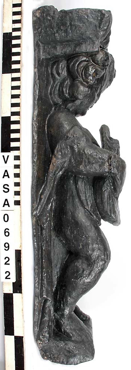 Skulptur föreställande en barnfigur med en cister (ett stränginstrument som tillhör familjen lutor).

Instrumentet hålls med bägge händer framför kroppen. Huvudet något vridet åt vänster. Kroppstyngden vilar på vänster ben medan höger ben är böjt. Utmed höger sida skymtar en mantel. Figuren står på ett minimalt underlag som troligen avser att föreställa ett stycke naturlig mark. Skulpturens baksida är slät.

Skulpturen är välbevarad.



Text in English: This is a sculpture of a child playing a cister.

It holds the instrument in both hands in front of its body. The head is turned slightly to the left. The weight of the body rests on the left leg, the right leg is bent. A mantle or cloak is to be seen on the figures right side. The child stands upon a small flat plinth. The back of the sculpture is smooth.

The sculpture is well preserved.