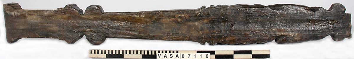 Skulpterad pilaster med övre delen i form av en karyatid med yppig, konstrik frisyr.
Höger arm hålls böjd med handen placerad framför bröstet. Vänster arm hålls utsträckt utefter sidan med handen vilande mot höften. Underkroppen döljs av ett hölje som framtill är prytt med stora, spiralformade ornament och små djurfigurer.
Karyatiden avgränsas från det rullverksornerade pilasterskaftet av en profilerad list. Nedtill avslutas skaftet av ett rullverksornament ovanför den rikt profilerade basen. Nedtill är pilastern försedd med en hög piedestal, prydd med beslagsornamentik. Figuren och dekoren omfattar även pilasterns sidor. Baksidan är slät.


Text in English: Sculpted pilaster with its upper part shaped as a Caryatid with a luxuriant artistic coiffure.
The right arm is held bent with the hand in front of the breast, the left arm is held at the side with the hand resting against the thigh. The lower part of the body is concealed by a shift or slip that is decorated with large spiral-shaped ornaments and small figures of animals.
A profiled batten separates the Caryatid from the shaft of the pilaster, which is ornamented with scrollwork. The shaft terminates with a scrollwork ornament above the richly profiled base. Below, the pilster is equipped with a high pedestal, decked with applied ornamentation. The back is smooth.