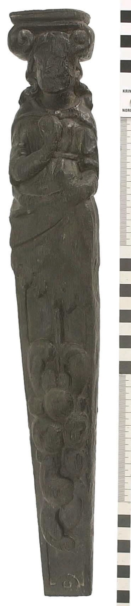 Hermpilaster.

Liten pilaster i form av en karyatid, med långt böljande hår under joniskt kapitäl.

Huvudet är vridet en aning åt höger. Med vänster hand hålls manteln samlad. I höger hand håller figuren en frukt eller dylikt framför bröstet. Pilasterskaftet är prytt med stora frukter och blad, fasthängande vid en stängel som delvis täcker det nedåt avsmalnande skaftets ramverk. Baksidan är slät.

Skulpturen är välbevarad, förutom ansiktet som är kraftigt slitet.

Fyra stycken spikhål, en övertäckt med en plugg.


Text in English: Sculpted small pilaster with a Caryatid with long wavy hair beneath an Ionic capital.

Its head is turned slightly to the right, its left hand holds its gathered mantle. Its right hand holds fruit or the like in front of its brests. The shaft of the pilaster is decorated with fruit and large leaves hanging from a stalk, which partly cover the framing of the tapering shaft. Its back is flat.

The sculpture is somewhat worn, its facial expression has been obliterated.