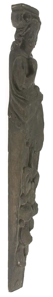 Hermpilaster.

Liten pilaster i form av en karyatid, med långt böljande hår under joniskt kapitäl.

Huvudet är vridet en aning åt höger. Med vänster hand hålls manteln samlad. I höger hand håller figuren en frukt eller dylikt framför bröstet. Pilasterskaftet är prytt med stora frukter och blad, fasthängande vid en stängel som delvis täcker det nedåt avsmalnande skaftets ramverk. Baksidan är slät.

Skulpturen är välbevarad, förutom ansiktet som är kraftigt slitet.

Fyra stycken spikhål, en övertäckt med en plugg.


Text in English: Sculpted small pilaster with a Caryatid with long wavy hair beneath an Ionic capital.

Its head is turned slightly to the right, its left hand holds its gathered mantle. Its right hand holds fruit or the like in front of its brests. The shaft of the pilaster is decorated with fruit and large leaves hanging from a stalk, which partly cover the framing of the tapering shaft. Its back is flat.

The sculpture is somewhat worn, its facial expression has been obliterated.