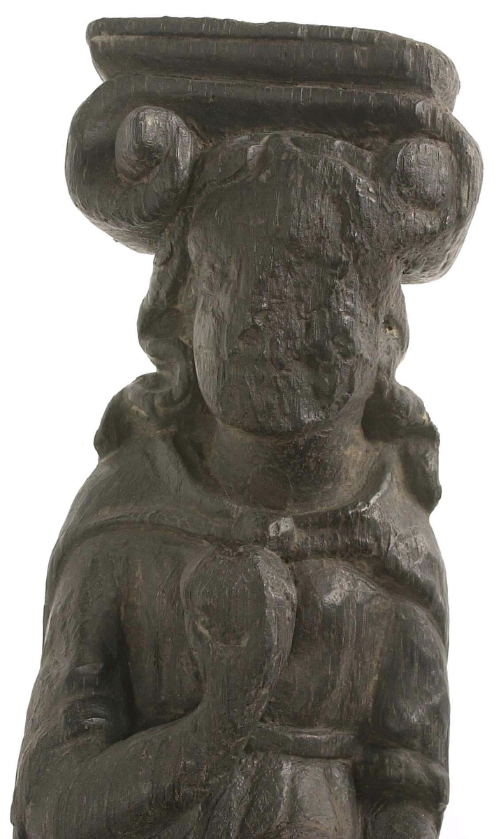 Hermpilaster.

Liten pilaster i form av en karyatid, med långt böljande hår under joniskt kapitäl.

Huvudet är vridet en aning åt höger. Med vänster hand hålls manteln samlad. I höger hand håller figuren en frukt eller dylikt framför bröstet. Pilasterskaftet är prytt med stora frukter och blad, fasthängande vid en stängel som delvis täcker det nedåt avsmalnande skaftets ramverk. Baksidan är slät.

Skulpturen är välbevarad, förutom ansiktet som är kraftigt slitet.

Fyra stycken spikhål, en övertäckt med en plugg.


Text in English: Sculpted small pilaster with a Caryatid with long wavy hair beneath an Ionic capital.

Its head is turned slightly to the right, its left hand holds its gathered mantle. Its right hand holds fruit or the like in front of its brests. The shaft of the pilaster is decorated with fruit and large leaves hanging from a stalk, which partly cover the framing of the tapering shaft. Its back is flat.

The sculpture is somewhat worn, its facial expression has been obliterated.