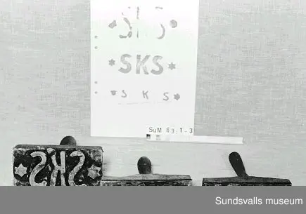 Stämpel i gummi med handtag i trä. Stämpeln visar bokstäverna SKS. Röda färgrester finns kvar på stämpeln.