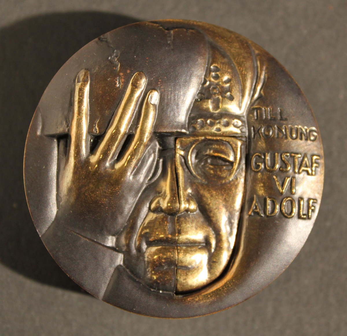 Medalj i två delar av patinerad brons. Åtsidans
yttersidavisar ett halvporträtt av kung Gustaf VI Adolf. Till vänster
synskungens hand hålla en urna, med text enligt MRK. Åtsidans
insidavisar den unge prins Gustaf Adolfs halvporträtt. Texten upptar
ettantal platser där utgrävningar skett i närvaro av eller på
initiativfrån kungen. Frånsidans yttersida visar nederst kungen inför
enutgrävning. Över hela motivet framtonar en kvinnokropp
symboliserandeModer Jord. Frånsidans insida visar ett tvådelat
ansiktsporträtt avden unge prinsen i nedsänkt relief till vänster och
Gustaf VI Adolf iupphöjd relief till höger, med text "Svenska
Arkeologiska SamfundetsHöge Beskyddare". Medaljen firar kungens 75 år
som arkeolog. Den gavsut av Svenska Arkeologiska Samfundet 1973. På
randen står medaljenslöpande tillverkningsnummer och tillverkaren
Sporrong instansat.