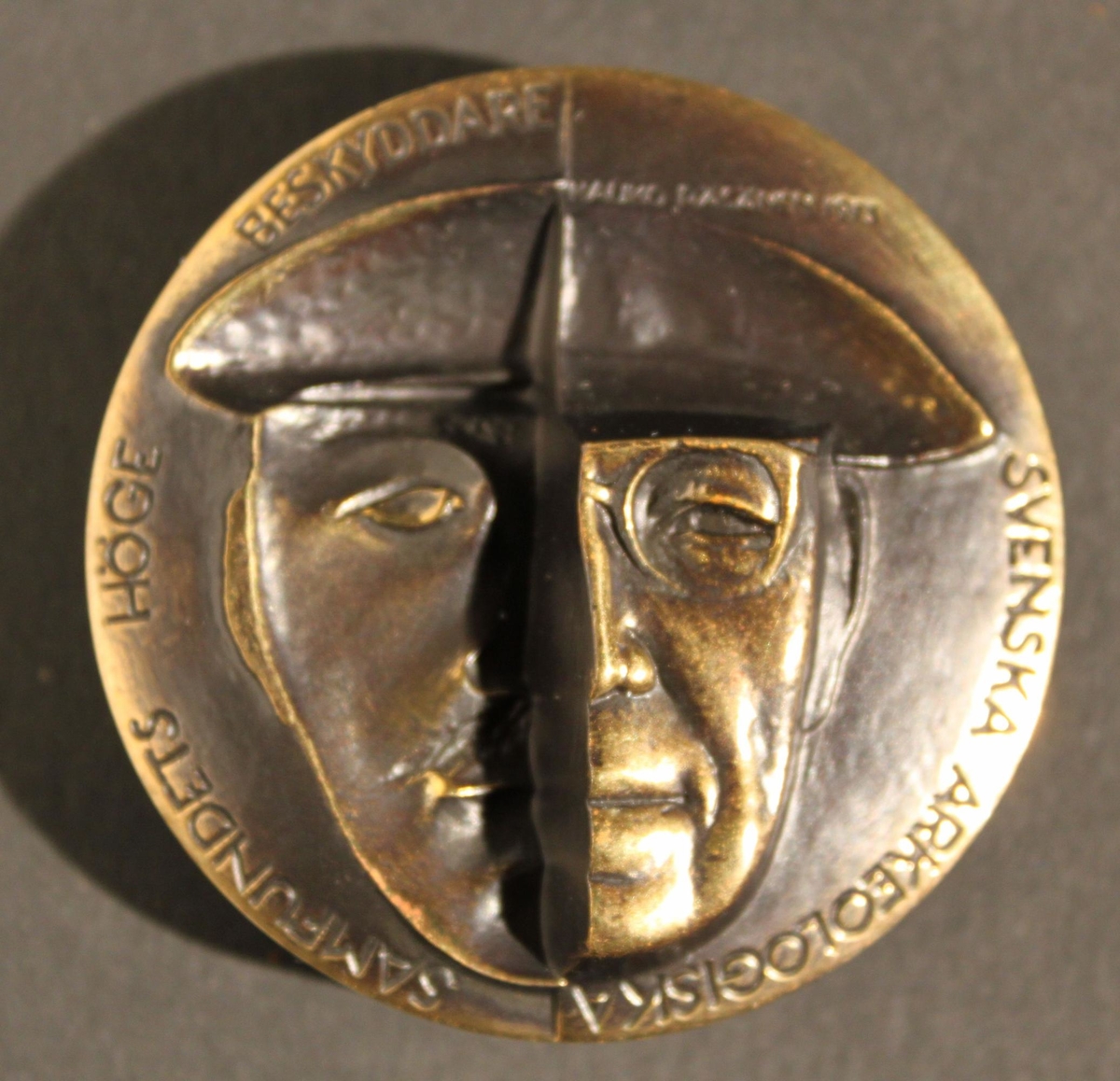 Medalj i två delar av patinerad brons. Åtsidans
yttersidavisar ett halvporträtt av kung Gustaf VI Adolf. Till vänster
synskungens hand hålla en urna, med text enligt MRK. Åtsidans
insidavisar den unge prins Gustaf Adolfs halvporträtt. Texten upptar
ettantal platser där utgrävningar skett i närvaro av eller på
initiativfrån kungen. Frånsidans yttersida visar nederst kungen inför
enutgrävning. Över hela motivet framtonar en kvinnokropp
symboliserandeModer Jord. Frånsidans insida visar ett tvådelat
ansiktsporträtt avden unge prinsen i nedsänkt relief till vänster och
Gustaf VI Adolf iupphöjd relief till höger, med text "Svenska
Arkeologiska SamfundetsHöge Beskyddare". Medaljen firar kungens 75 år
som arkeolog. Den gavsut av Svenska Arkeologiska Samfundet 1973. På
randen står medaljenslöpande tillverkningsnummer och tillverkaren
Sporrong instansat.