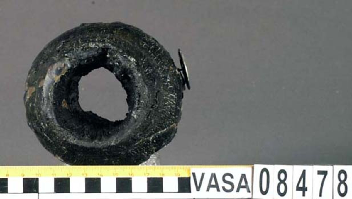 Kolv till ansättare. Kolven består av ett cylindriskt svarvat trästycke med ett centrerat hål för skaftet. Dess övre ände, mot skaftet till, har ursprungligen varit profilsvarvad. Kolven är kraftigt eroderad varför profilsvarvningen är delvis utplånad.
