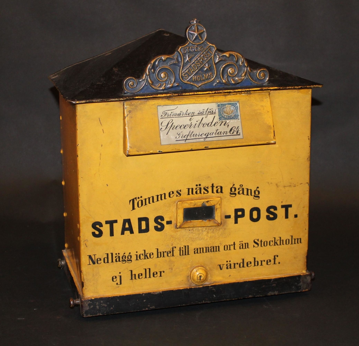 Brevlåda med sluttande, brutet tak. I främre delen av taket ett av Stockholms stadspost emblem. Brevlådans tak och ramen runt brevlådans botten är svart i övrigt är brevlådan gul. På inläggsklaffen ett vitt kuvert med ett av Stadspostens frimärken och texten "Frimärken säljas i Speceriboden Grefturegatan 64".
Där under en liten glasruta avsedd för visning av tömningstider. Brevlådans front har inskription. Brevlådan tömmes från botten med en tömningsväska. Ett lås är centrerat i lådans nederkant. Nyckeln har PM 1642.