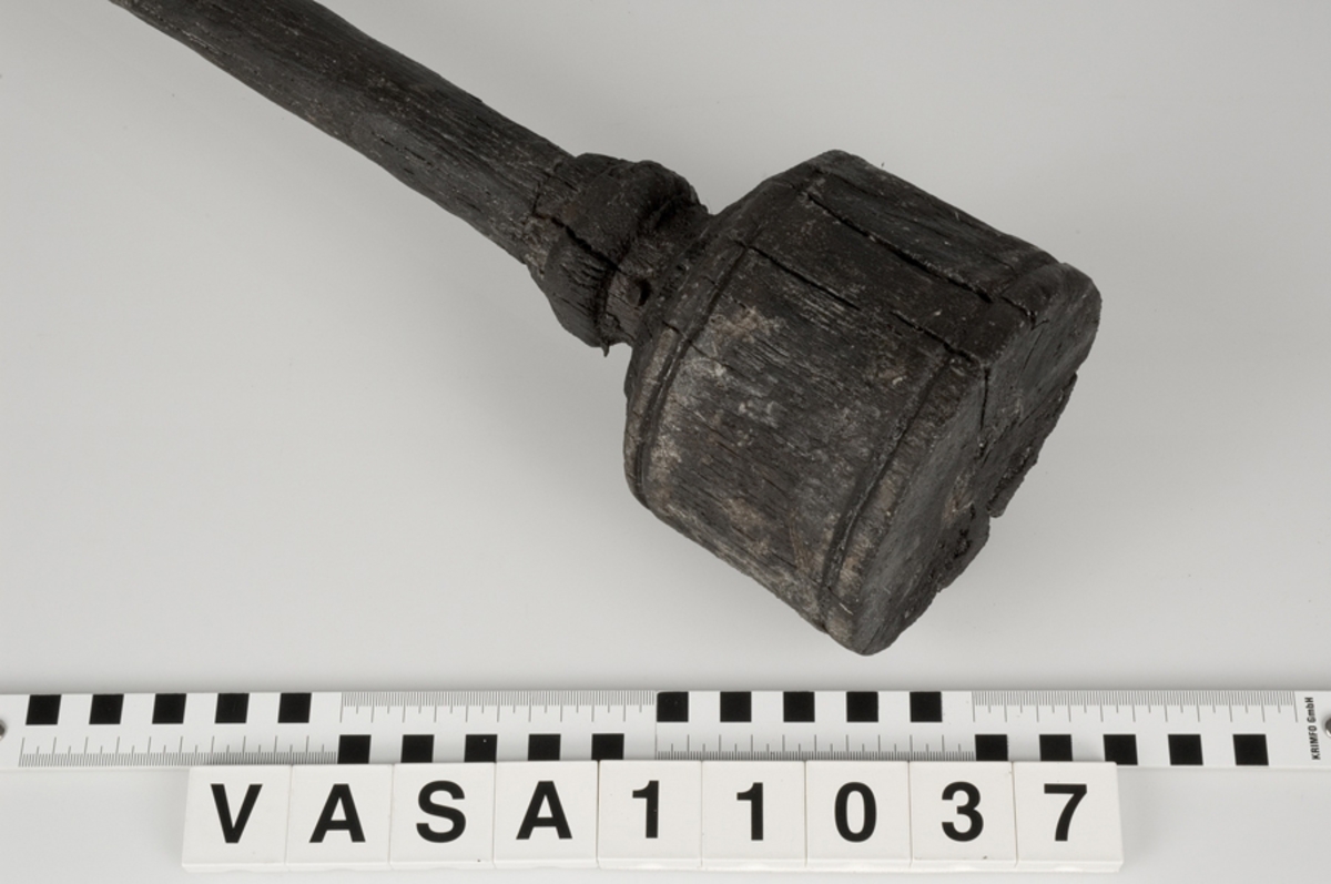 Kolv till ansättare. Kolven består av ett cylindriskt, svarvat, trästycke med ett centrerat skafthål. I övre änden, åt skaftet till, är kolven profilsvarvad. Motsatt ände är platt. Några sprickor, annars i fint skick.