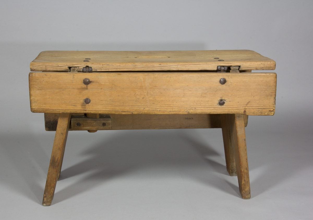 Pall eller pallbol i furu. Låg, avlång med bockformade ben. På pallens ena långsida väl tilltagen sarg, på andra sidan av fälltyp, fäst med gångjärn. Handtag i sätet i form av ett hål.