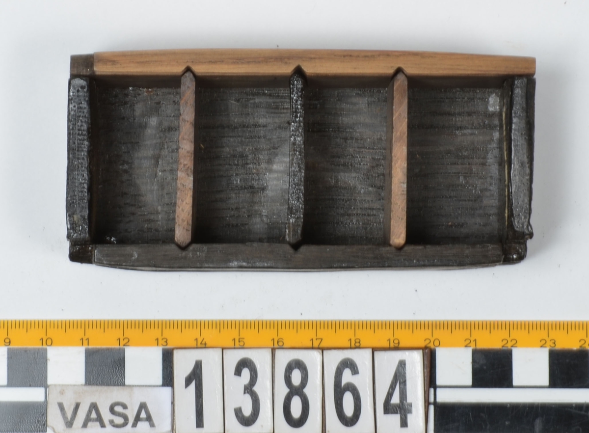 Ett litet träskrin med skjutlock.
Skrinets gavlar och långsidor är sinkade. Bottnen sitter infälld i gavlarna och är fästad med trästift vid långsidorna. Skrinet är försett med tre, löstagbara mellanväggar. På insidan av skrinet finns spår för mellanväggarna och för locket. Lockets ena ände är profilerad och har en liten halvcirkelformad urholkning som fungerar som tumgrepp. Den profilerade änden skjuter ut en bit utanför skrinet, då locket är stängt.
Två av skrinets mellanväggar samt en av långsidorna är rekonstruerade. Längs med skrinets ena långsidan löper en spricka. Skrinet är i gott skick.